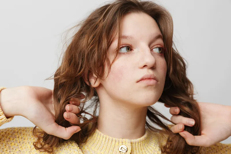 Young woman with curly hair wearing yellow sweater looks away thoughtfully. Close-up portrait with natural light on white background. Concept of contemplation and casual style.
