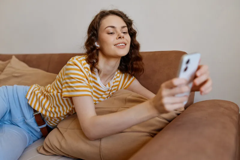 Young woman lounges on a cozy living room sofa, taking a casual selfie with her smartphone, feeling relaxed and confident at home.