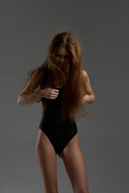 Young woman in black swimsuit with long hair posing on gray background, soft lighting, thoughtful mood