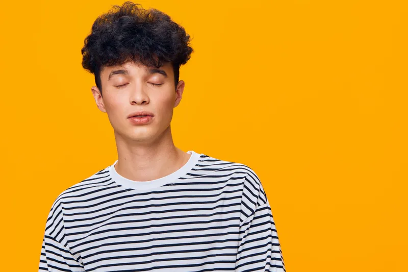 young man with closed eyes wearing striped shirt stands peacefully against vibrant orange background, expressing calmness and introspection, casual fashion style