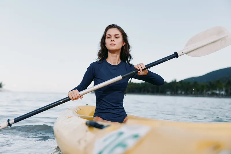 A focused female kayaker paddles a yellow kayak across calm water, wearing a dark long sleeve top, arms extended, capturing strength, balance, and motion on a sunny day.