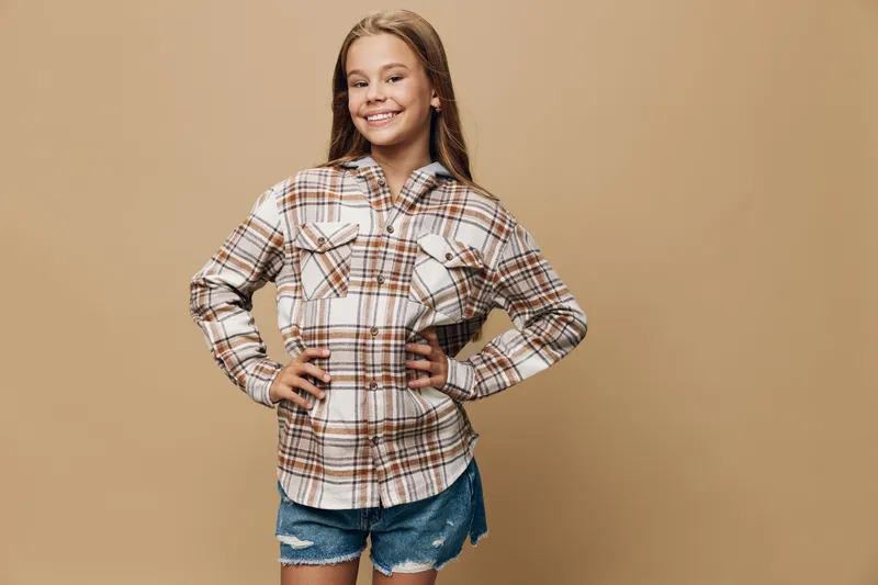 Teenage girl smiling with hands on hips wearing plaid shirt and denim shorts standing against plain beige background, confident and casual look.