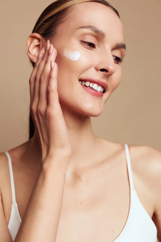 Young woman with clear skin applies moisturizing cream on cheek smiling gently wearing white tank top isolated on beige background skincare beauty routine concept.