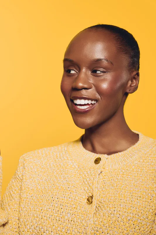 Smiling African woman with dark skin wearing a yellow knitted sweater looks away with a bright cheerful expression. Studio portrait on a yellow background. Concept of happiness and beauty.