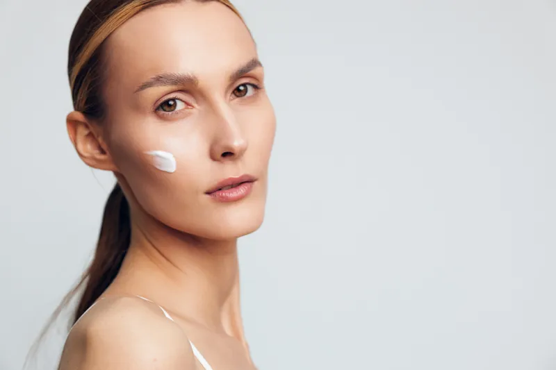 Young woman with long blonde hair applies white face cream on her cheek while looking at the camera against a plain light background.