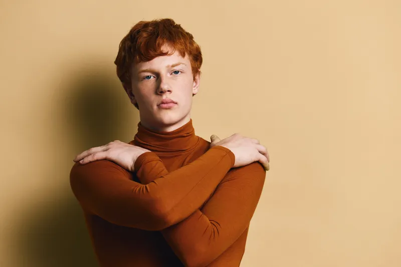 Portrait of young man with red hair wearing brown turtleneck sweater posing with crossed arms on beige background with soft shadow and serious facial expression.