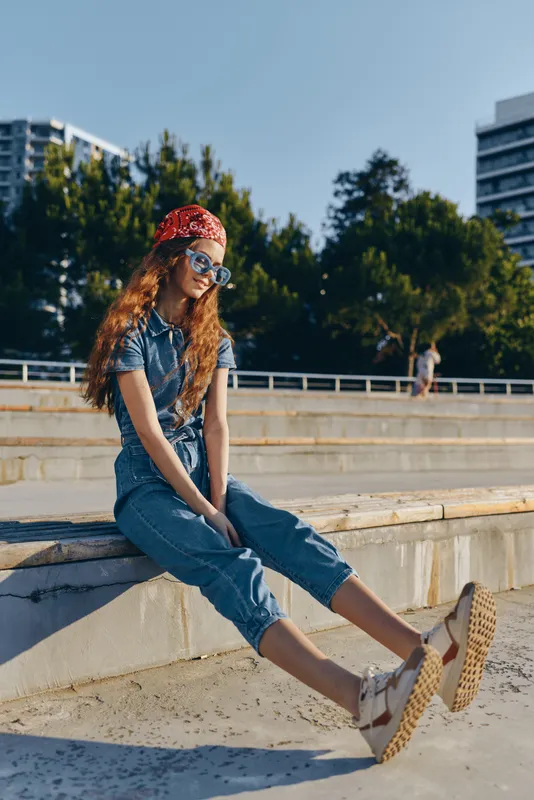 redhead woman wearing denim jumpsuit and bandana sits outdoors on sunny day with sunglasses and casual shoes near urban park under blue sky