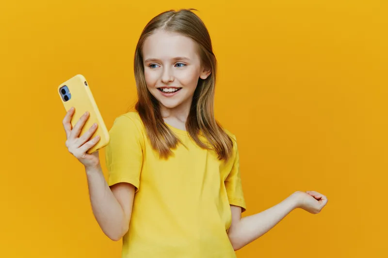 Cheerful girl in yellow shirt holding smartphone on yellow background, expressing joy and excitement while taking a selfie