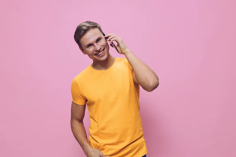 Smiling young man in bright yellow t-shirt talking on smartphone while standing against pink background. Casual style, cheerful expression, positive mood, lifestyle concept, studio portrait, young adu