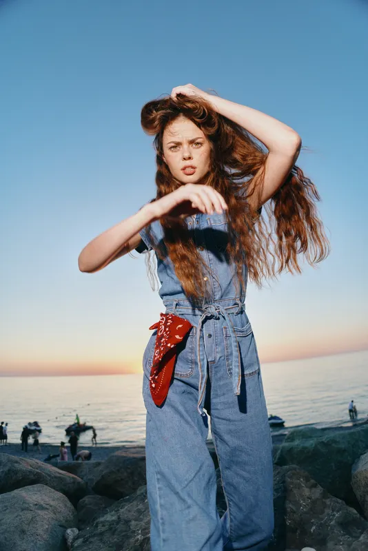 Lifestyle woman in modern boho-western denim street style posing outdoors by the rocky beach during sunset with film color tones and natural background.