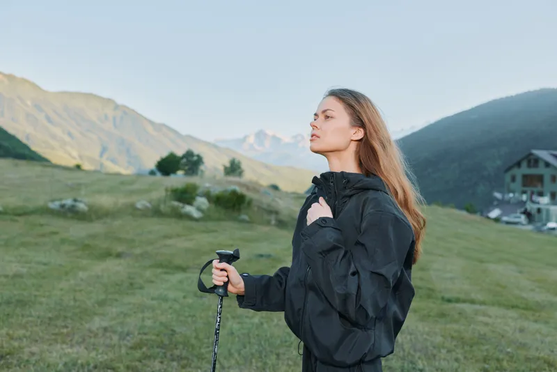 A lone woman stands outdoors in a rugged mountain landscape, wearing a dark jacket and gripping a trekking pole. She gazes upward with calm, focused poise.