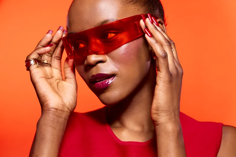 Stylish black woman with dark skin wearing red sunglasses and a red top poses confidently against an orange background. Concept of fashion, beauty, and modern style.