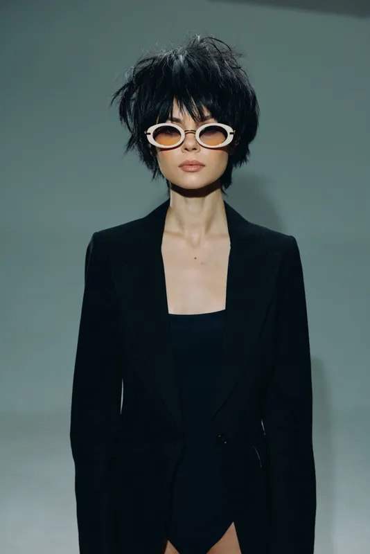 Stylish woman wearing white sunglasses and black blazer with short curly hair in studio setting