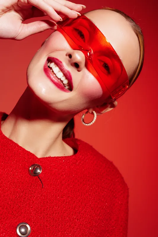 Smiling woman wearing red transparent glasses and red sweater poses against red background. Close-up portrait with hoop earrings and bright makeup. Concept of fashion, style, and vibrant mood.