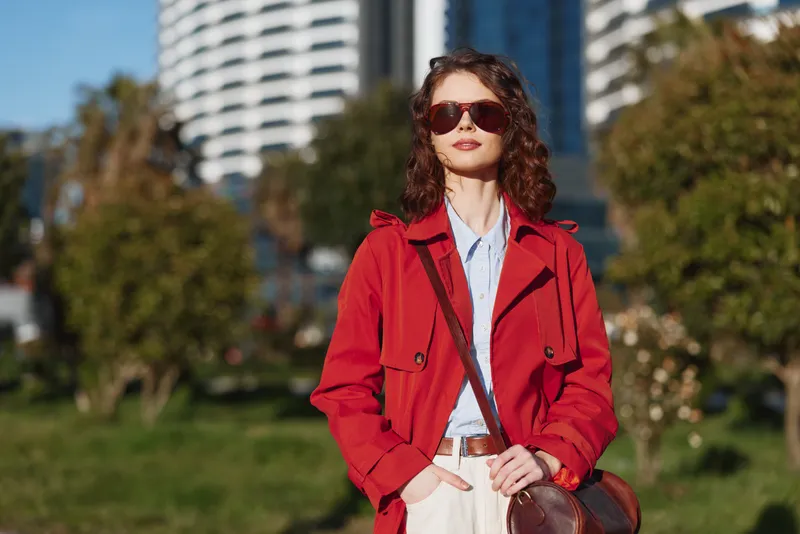 Stylish young woman in a red trench coat stands confidently in a sunlit park with modern buildings behind, posing with a bag and sunglasses, and a relaxed smile.