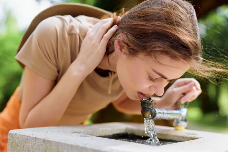 Woman drinking water from a concrete fountain in an outdoor park during summer, young female hydrating in casual clothes, closeup profile showing refreshment and natural sunlight.