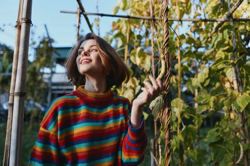 Woman in a colorful striped sweater smiling in an urban garden, looking up with short hair, young adult enjoying warm sunlight while touching vines in a cozy lifestyle fashion portrait.