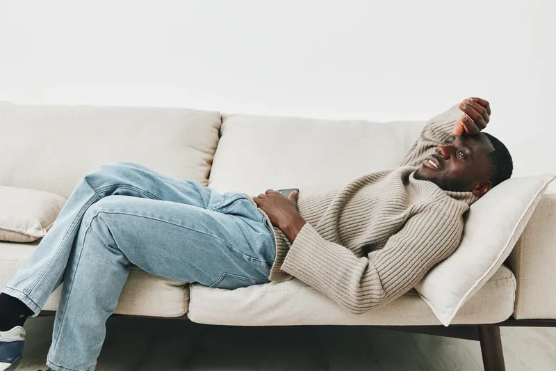 Young Black man in casual attire relaxed on a sofa, expressing emotions of stress and discomfort while holding his head