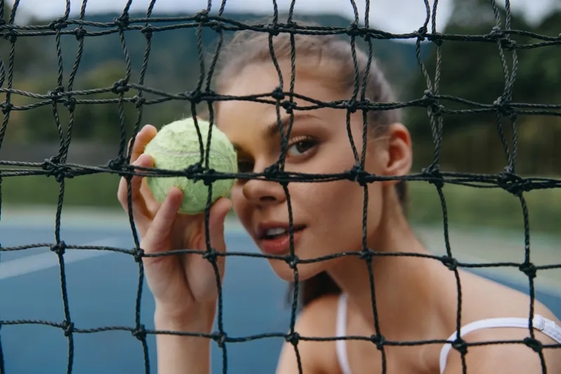 Young female athlete holding a tennis ball, looking through a tennis net on a court, showcasing determination and focus during practice in a natural setting