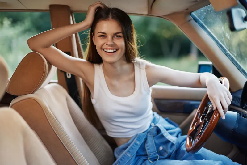 Happy young woman in casual outfit smiling in vintage car, showcasing carefree attitude and youthful energy in outdoor setting with natural light.