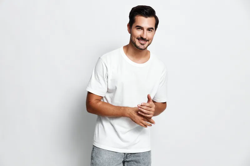 Smiling young man wearing a white t-shirt poses with hands clasped, exuding warmth and approachability against a clean, neutral background. People lifestyle concept.