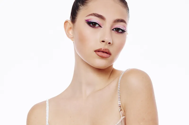 Young woman with creative fashion makeup poses against white background. She features long lashes, glossy lips, and elegant fashion clothing with thin straps for beauty editorial concept.