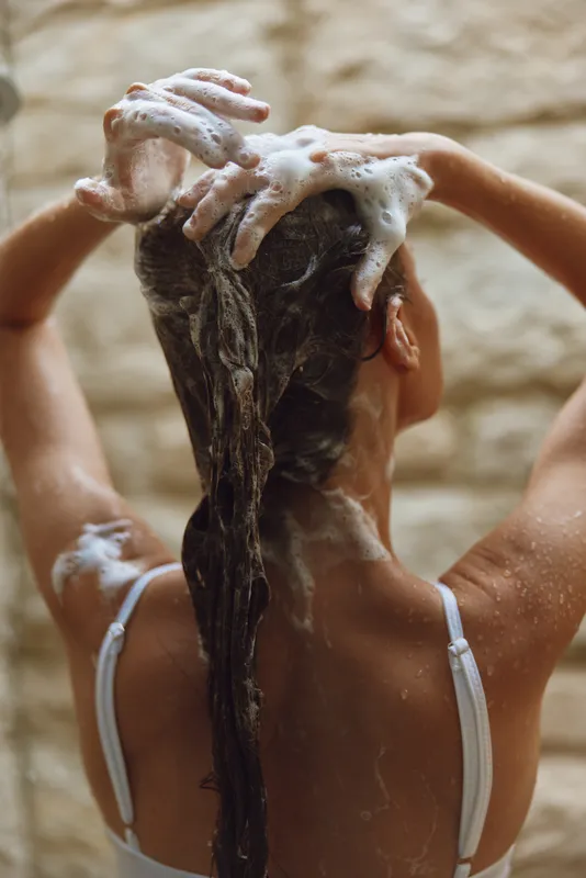 Woman washing long hair with shampoo foam, showing back view and wet strands in natural indoor bathroom setting, emphasizing self-care and cleanliness.