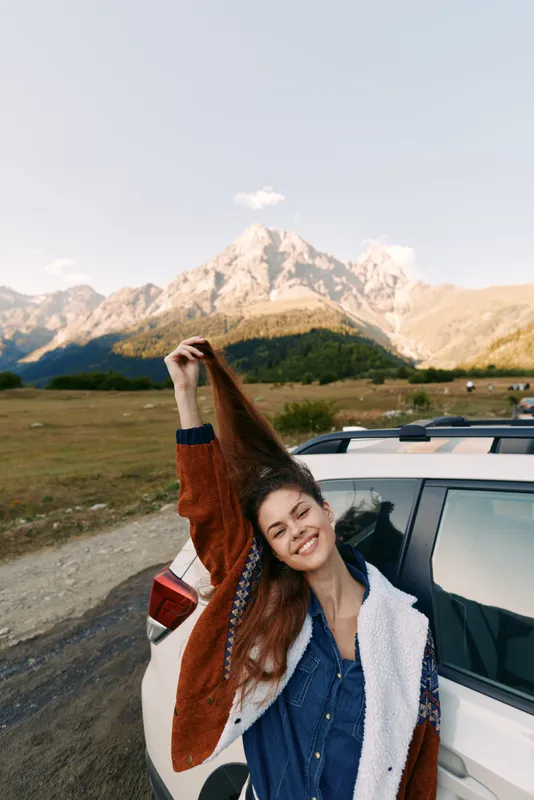 Woman car mountain road outdoors travel portrait happy smiling near vehicle at scenic roadside, young female on adventure with cozy jacket, nature landscape and freedom vibe.