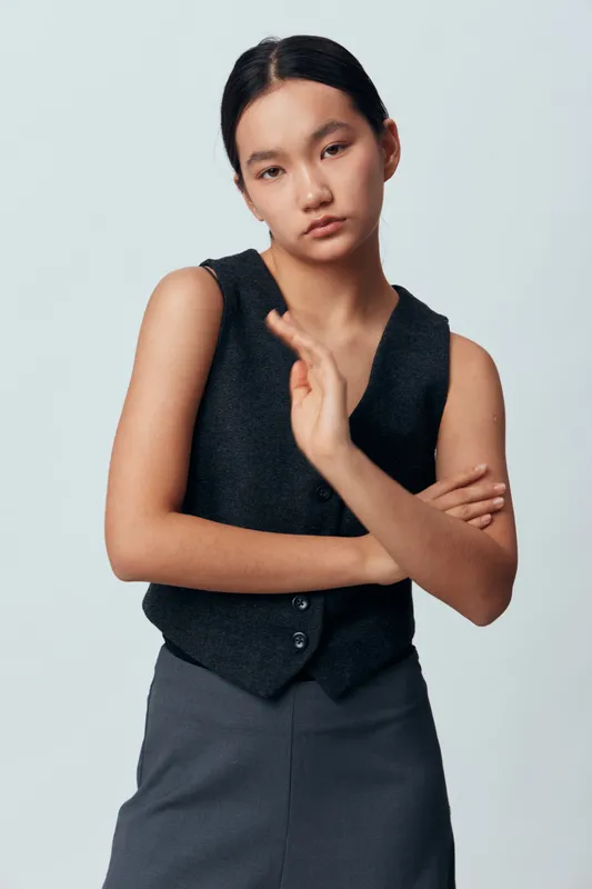 Young woman in black vest and gray skirt poses with crossed arms and serious expression against light background. Casual fashion, portrait, standing and confident female model.