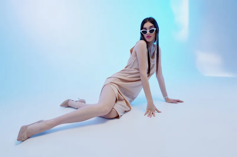 Fashionable woman in a beige dress and sunglasses posing on the floor in a studio with blue lighting. Concept of beauty, fashion, elegance, and modern style.