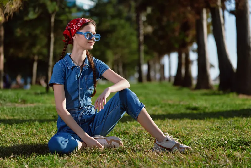 Young woman sitting on grass in denim outfit with sunglasses enjoying sunny day in park surrounded by trees and natural green environment.