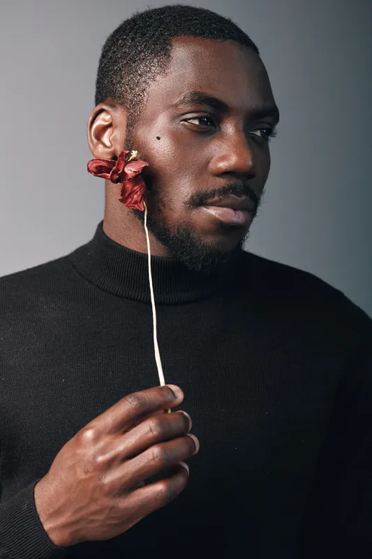 African man with dark skin holding dried flower near face in creative conceptual visual style. Portrait of thoughtful male model wearing black turtleneck with artistic expression and minimal