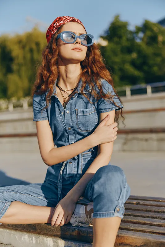 Lifestyle woman in modern boho-western denim street style wearing blue sunglasses and red bandana, relaxed outdoors with natural background in warm film color tones.