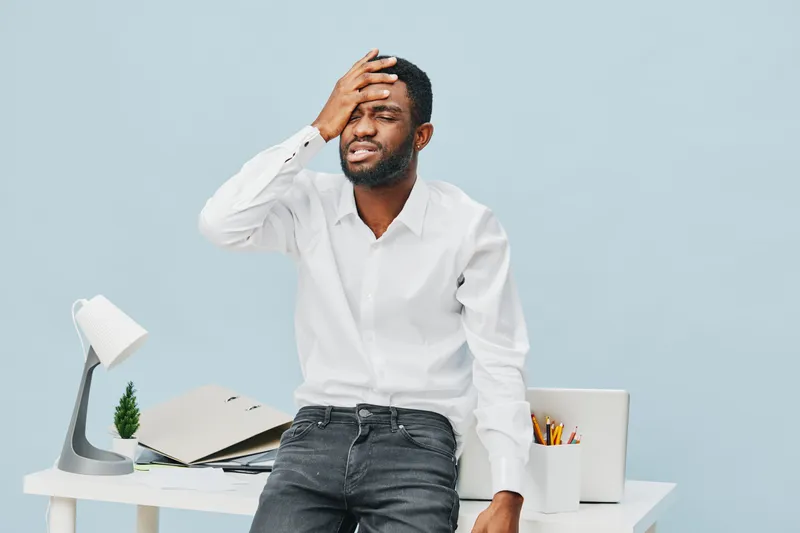 Frustrated young african american man in white shirt feeling stressed holding head in office with workspace background, office work pressure concept, workplace stress, emotional fatigue, tired profess