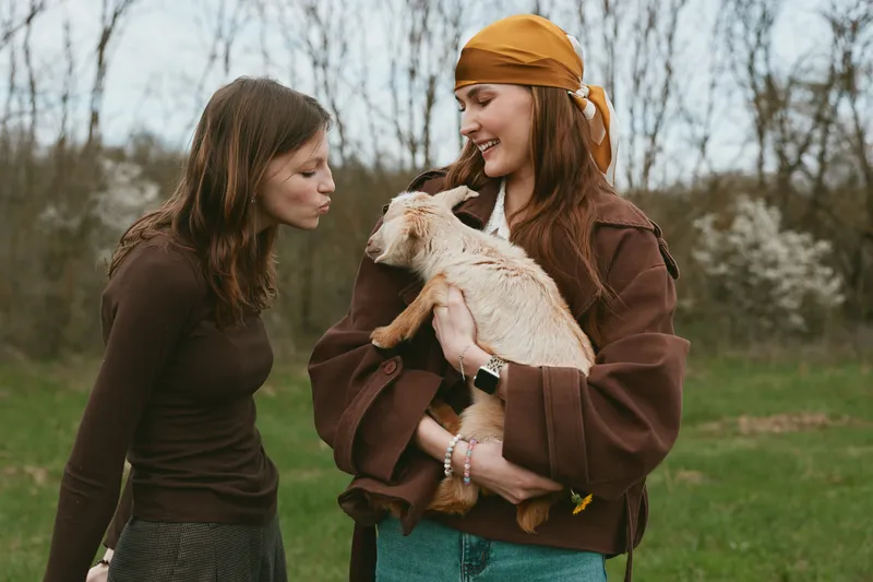 Two women in casual clothing interact with a playful goat kid outdoors, showing warm friendship, gentle affection, and rural charm amid a grassy field and bare trees.