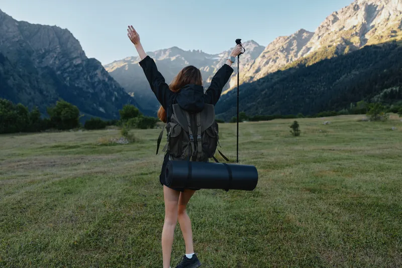 A lone hiker stands in a wide valley, arms raised in triumph as towering mountains rise behind. She wears a backpack and carries a rolled mat, ready for adventure.