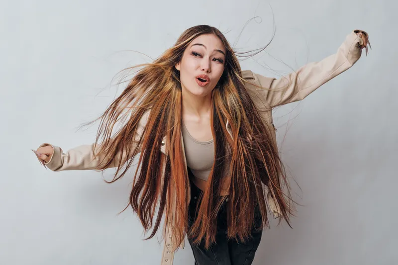 Woman with long braided hair wearing casual beige jacket and tank top posing energetically on light background. Fashionable female with vibrant hairstyle and expressive face.