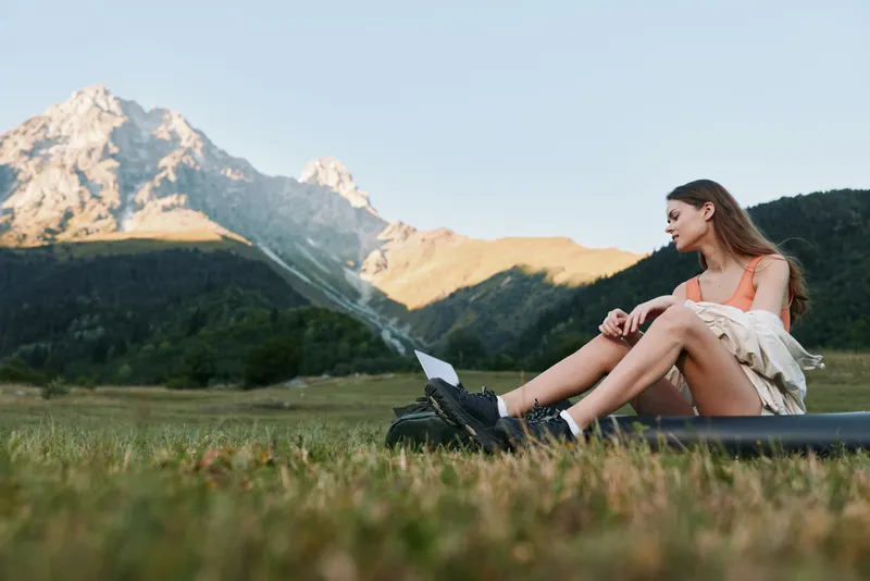 A thoughtful young woman sits on lush grass with a laptop, gazing toward distant mountains under a clear blue sky, creating a calm, productive outdoor work scene.