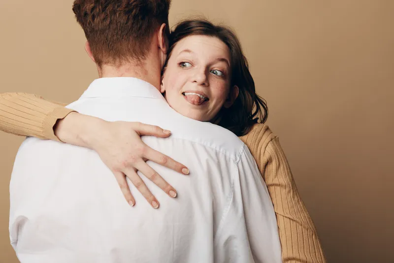 Young woman in beige sweater hugs man in white shirt from behind, playfully sticking out her tongue. Intimate moment with warm beige background, concept of affection and casual relationship.