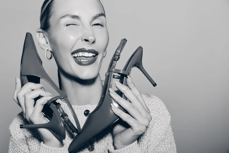 Happy white woman with fair skin holds two high heel shoes near face smiling joyfully in cozy sweater. Concept for fashion, style, footwear, and female lifestyle.