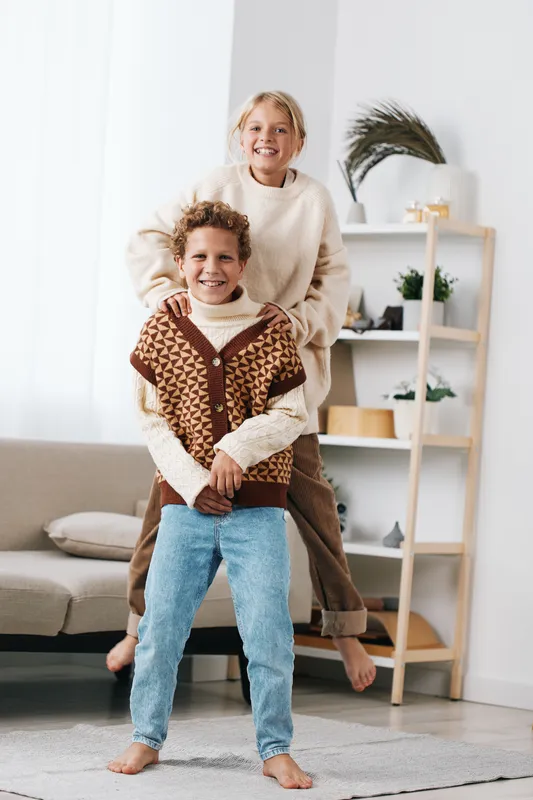 Two young siblings playfully pose indoors, one perched on the other's back, wearing cozy sweaters in a bright living room, radiating warmth, laughter, and cheerful energy.