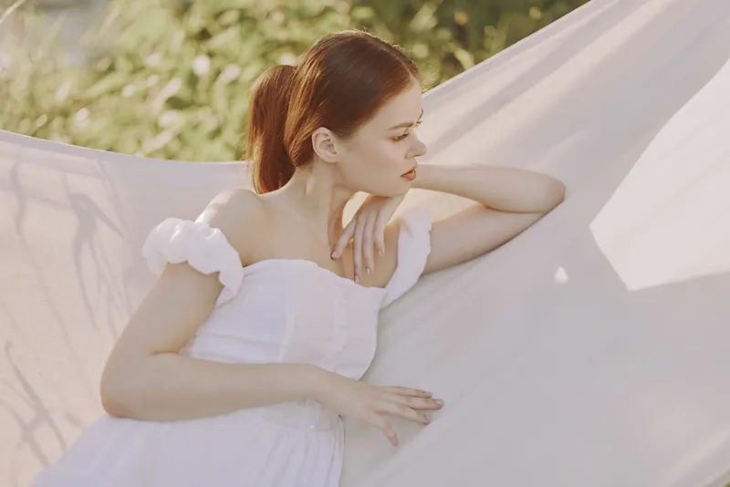 A serene young woman in a white dress relaxing in a hammock, surrounded by lush greenery, conveying a sense of calm and tranquility during a summer day