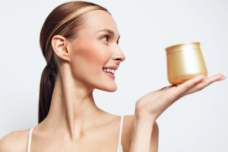 Smiling woman in a white top holds a gold cream jar up to eye level, celebrating skincare moment with soft studio light, clean backdrop, healthy glow, calm confidence and radiant skin tone