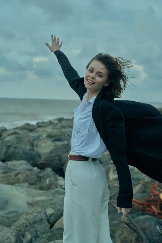 Confident businesswoman on a windy rocky shore, smiling at the camera, wearing a white blouse, dark blazer, and flowing coat, capturing motion and freedom in an outdoor workplace scene.