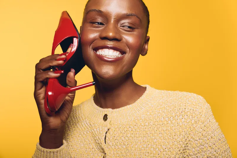 Smiling African woman with dark skin holds red high heel shoe near her face against yellow background. Concept for fashion, beauty, happiness, and stylish accessories.