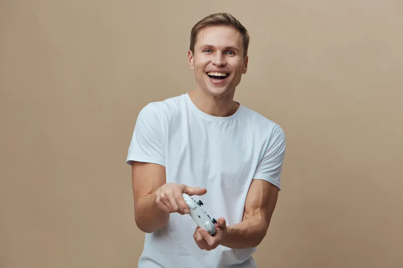 Happy young man holding a game controller smiling, casual clothing, beige background, joyful expression, indoor setting, user enjoyment, lifestyle concept, relaxed mood.