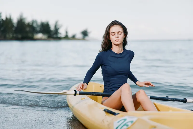 A young woman sits in a yellow kayak at the shoreline, paddling with a single oar, sunlit water surrounds her, creating a calm mood and sense of adventure.