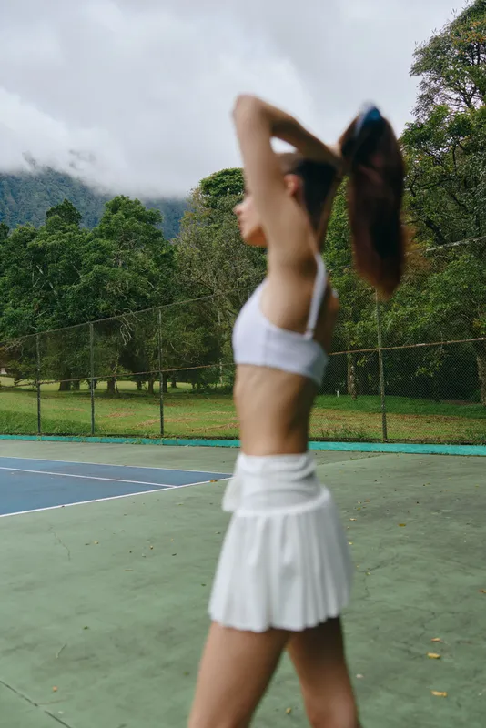 Young athletic woman stretching on a tennis court, wearing a white sports outfit, surrounded by lush green trees and a cloudy sky, showcasing health and fitness