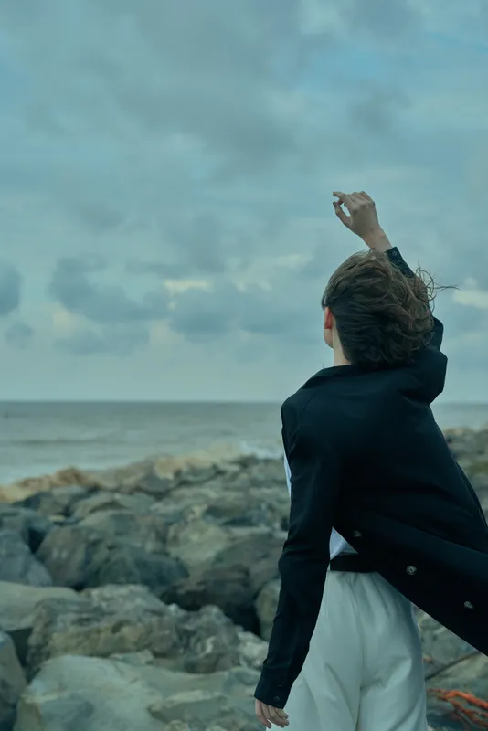 Woman in a dark jacket and white skirt stands on a rocky coastline, wind sweeping hair, reaching toward the cloudy horizon to convey motion, resilience, and outdoor adventure.