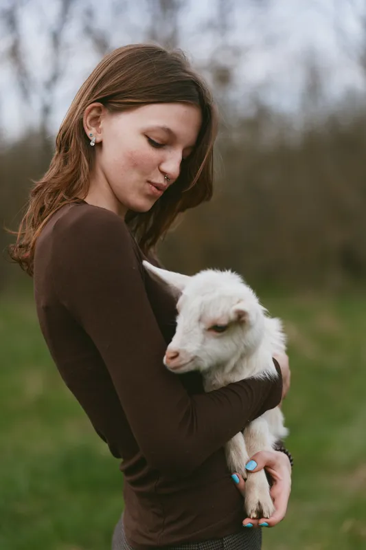 Cheerful woman outdoors, embracing a gentle goat kid, serene moment, soft light, earthy tones, lush meadow backdrop, candid lifestyle scene, natural connection with animals, peaceful mood, outdoor